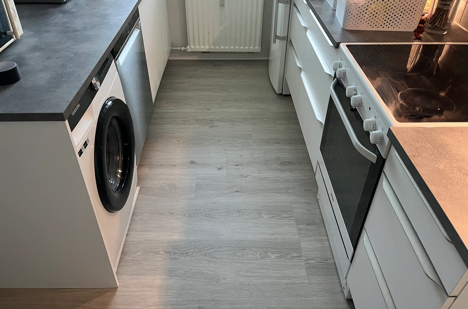 Lyst gråt klikgulv lagt gennem et smalt køkken mellem hvide skabe, mørk bordplade og hvid radiator
