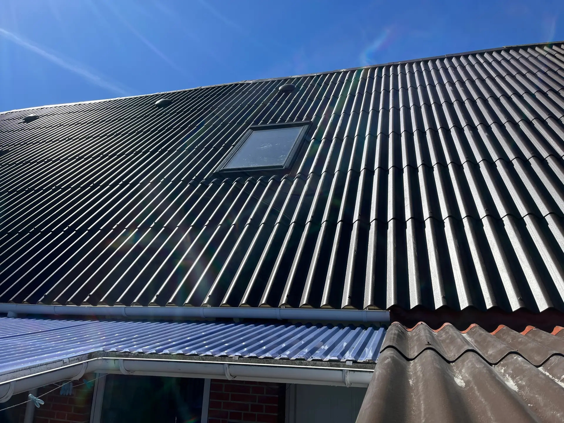 Nyt sort profiltag tæt på med ét ovenlysvindue, hvid tagrende og skarp blå sommerhimmel