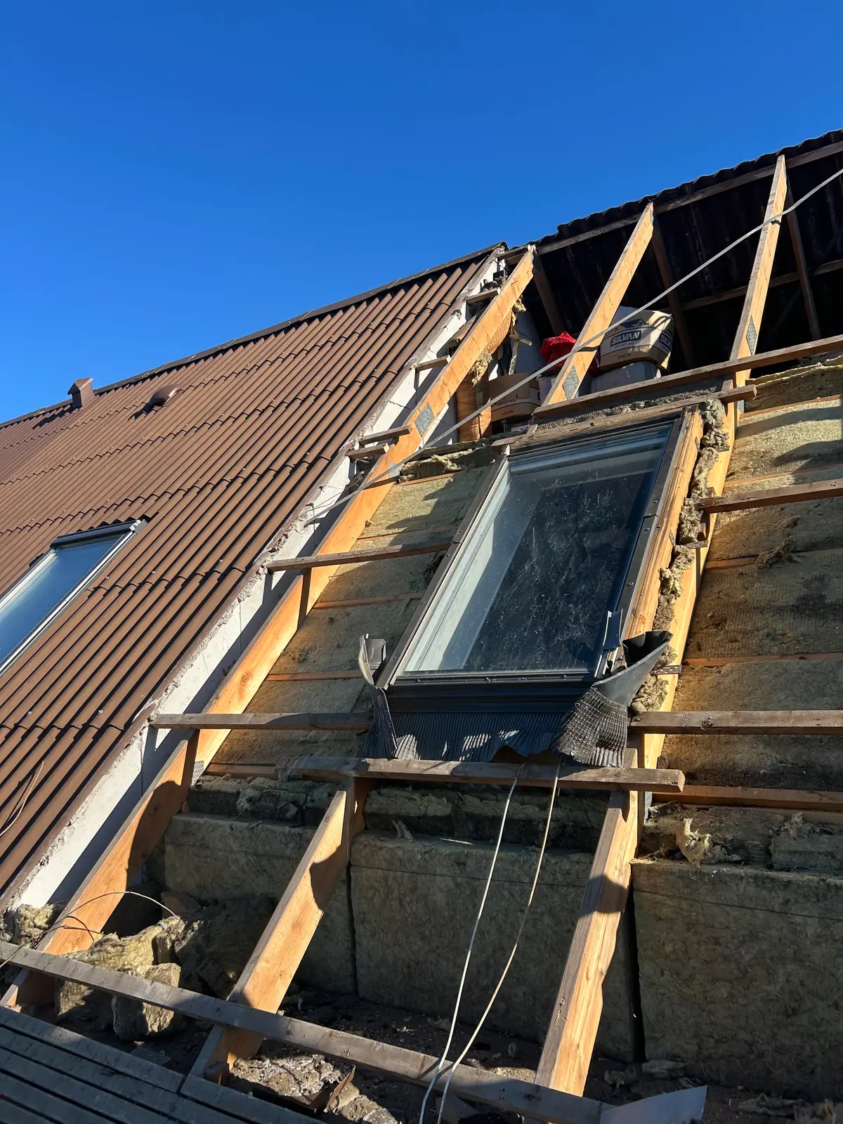 Tagflade under renovering med nyt ovenlysvindue, friske lægter, blotlagt mineraluld og gamle brune tegl mod blå himmel