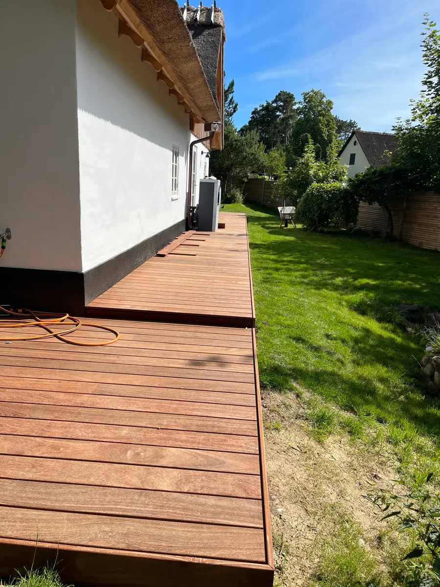 Ny rødbrun hårdttræsterrasse langs hele facaden på et hvidpudset stråtækt hus med sort sokkel, varmepumpe på muren, orange kabel på dækket og græsplæne ved siden af